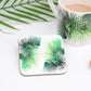A square coaster with a green leaf design, presented next to a cup of coffee and a wooden spoon, on a white background.