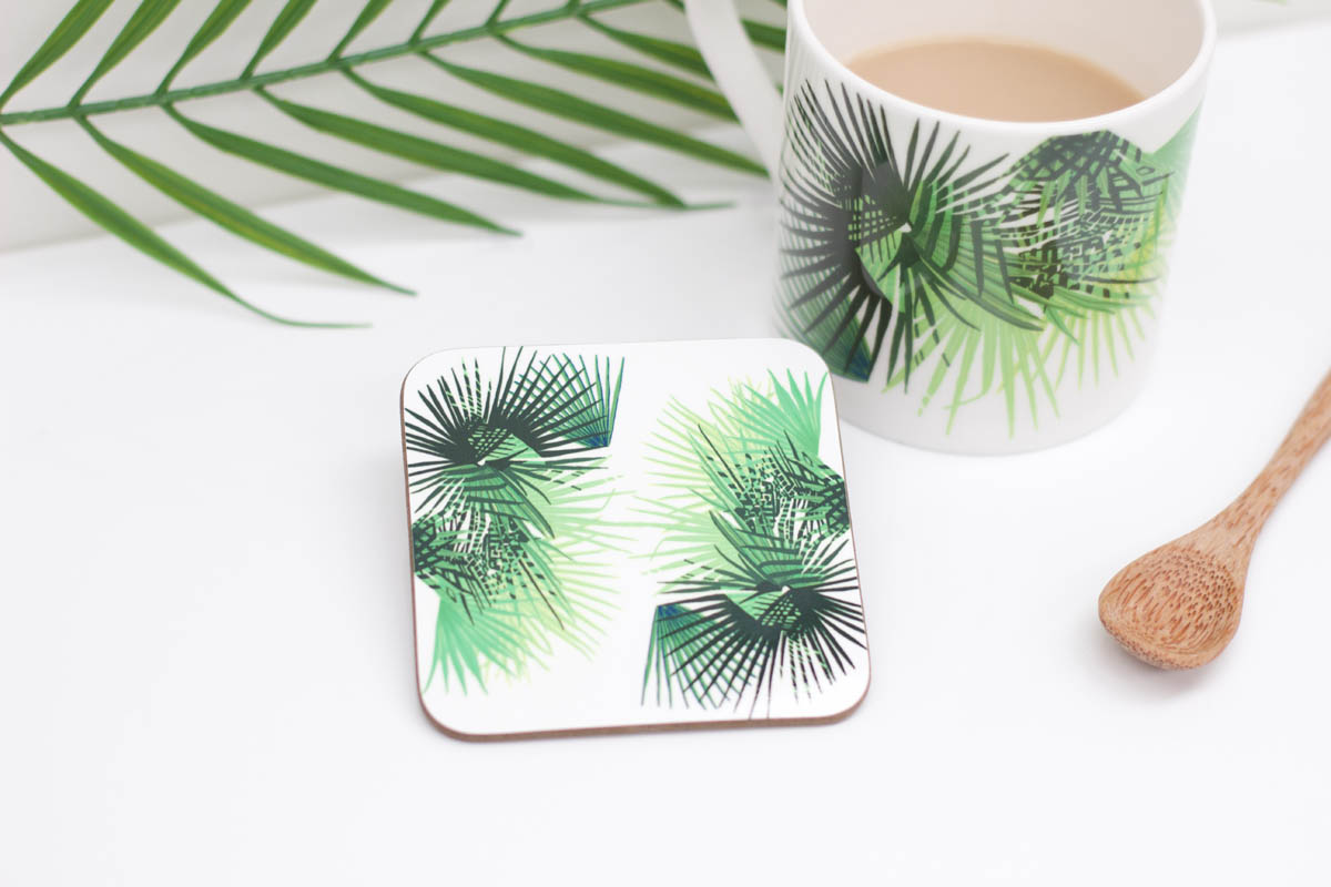 A square coaster with a green leaf design, presented next to a cup of coffee and a wooden spoon, on a white background.