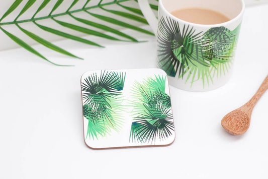 A square coaster with a green leaf design, presented next to a cup of coffee and a wooden spoon, on a white background.