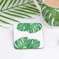 A square coaster with a tropical leaf design, specifically a cheese plant, on a white background. The coaster is placed on a white surface with a cup of tea and a wooden spoon in the background.