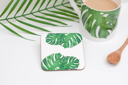 A square coaster with a tropical leaf design, specifically a cheese plant, on a white background. The coaster is placed on a white surface with a cup of tea and a wooden spoon in the background.