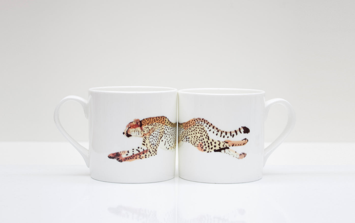 Two white bone china mugs with a cheetah design in the center, placed on a white surface.