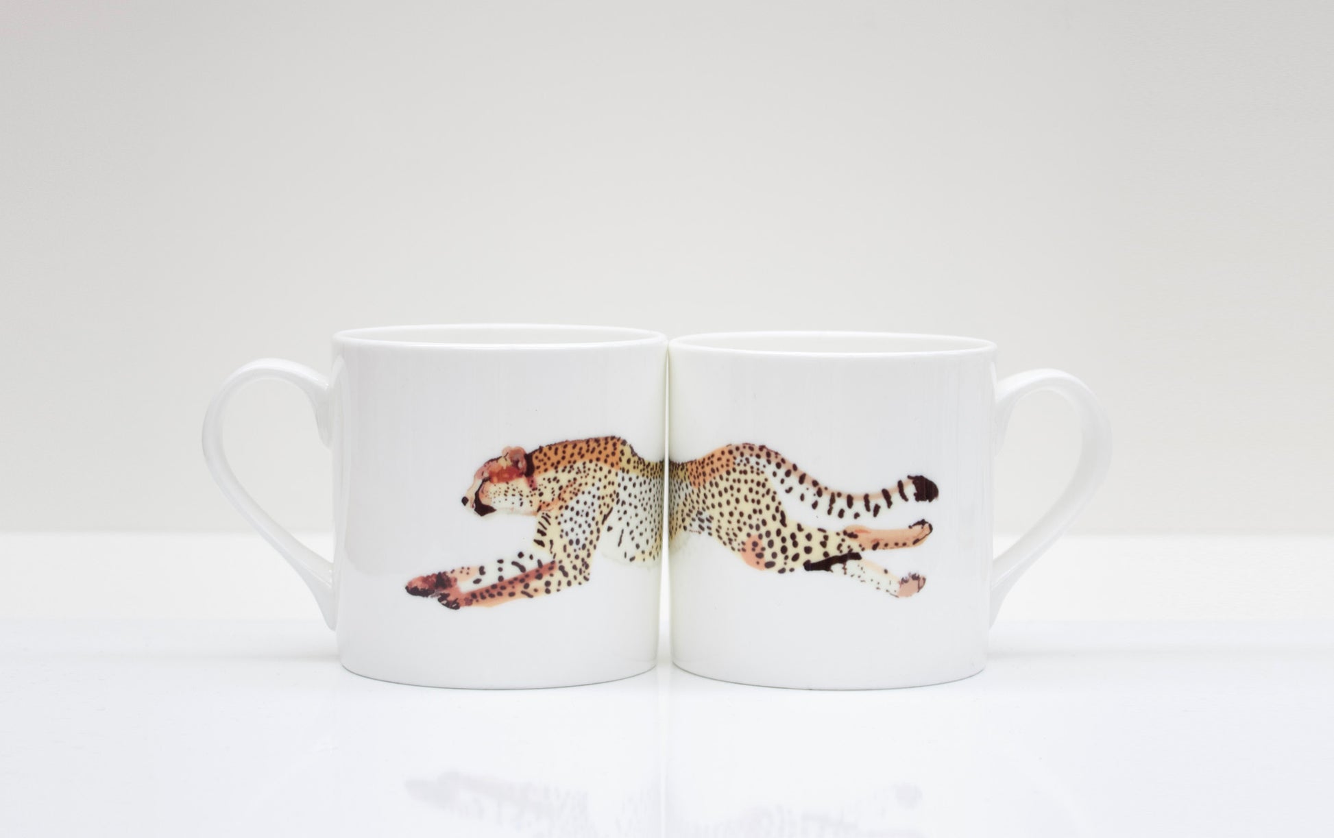 Two white bone china mugs with a cheetah design in the center, placed on a white surface.