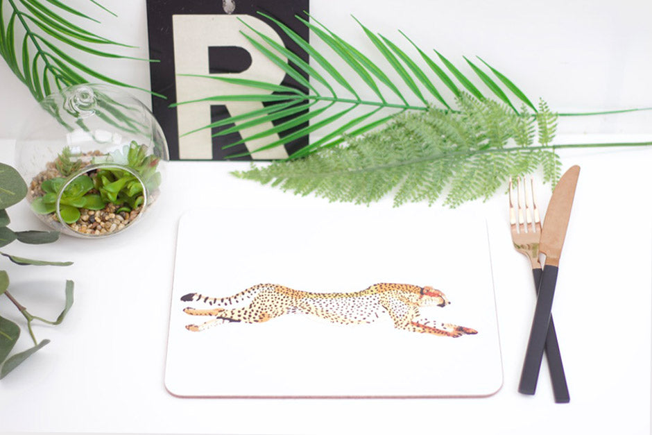 A white placemat with a drawn illustration of a cheetah, displayed on a table with cutlery and decorative plants around it.