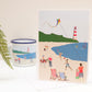 A greetings card with a colorful illustration of a beach scene, including a kite, people in swimwear, and a lighthouse, with a blue sky and green landscape in the background.