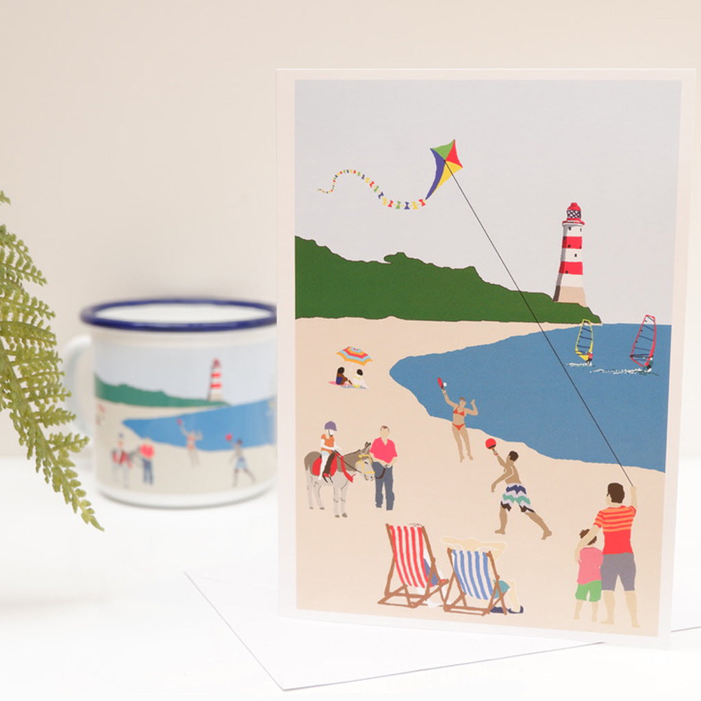 A greetings card with a colorful illustration of a beach scene, including a kite, people in swimwear, and a lighthouse, with a blue sky and green landscape in the background.
