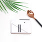 A square white coaster with a black geometric symbol design, displayed next to a copper-colored spoon on a white surface with a green leaf in the background.