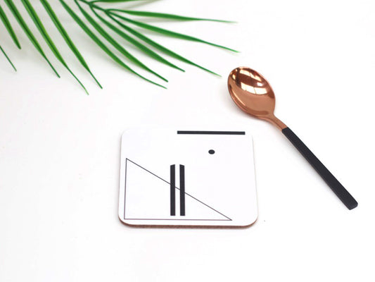 A square white coaster with a black geometric symbol design, displayed next to a copper-colored spoon on a white surface with a green leaf in the background.