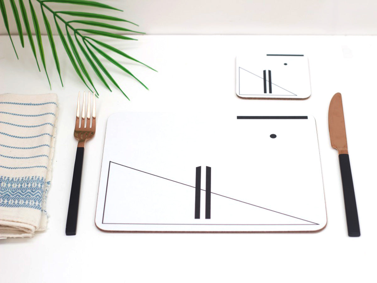 A placemat with which and black symbol designs, displayed on a table with a matching coaster, blue and white striped napkin, a brown fork, and a green palm leaf in the background.