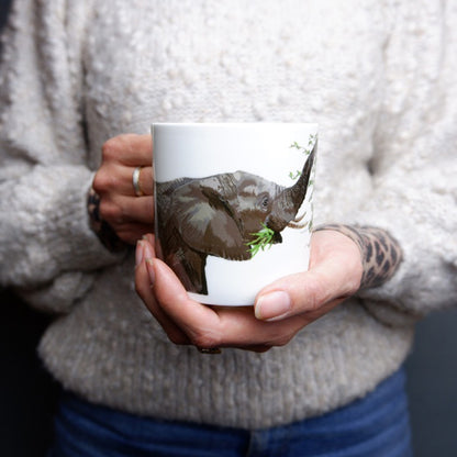 A person holding a white bone china mug with an illustration of an elephant reaching its trunk around the mug.