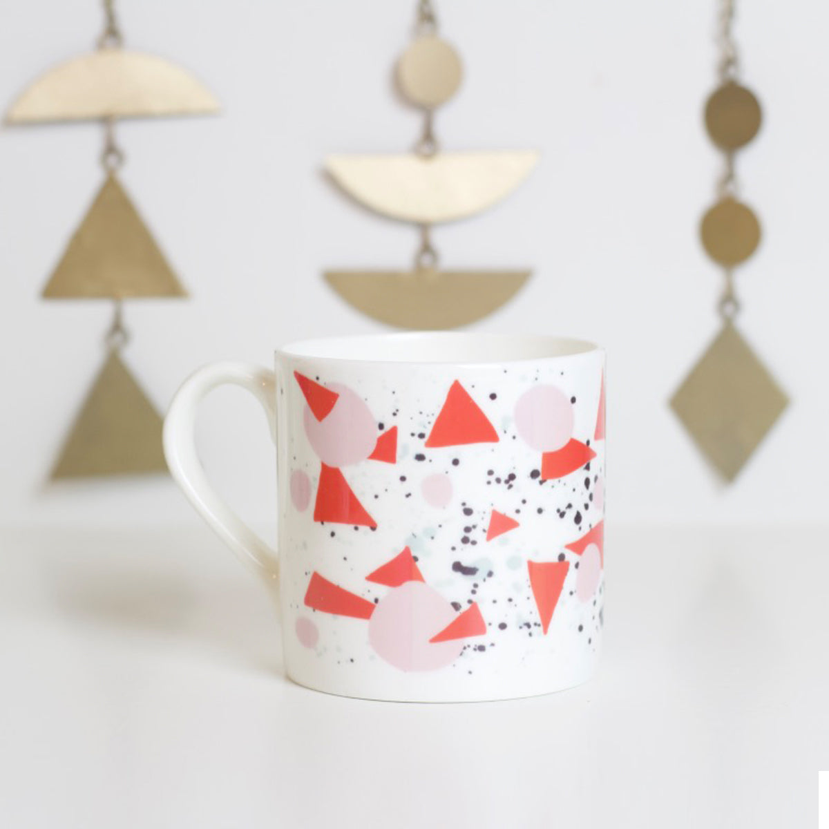 A white bone china mug with a pattern of bold red triangles, subtle pink curves, and black and grey splashes, displayed on a white surface with geometric metal decorations in the background.