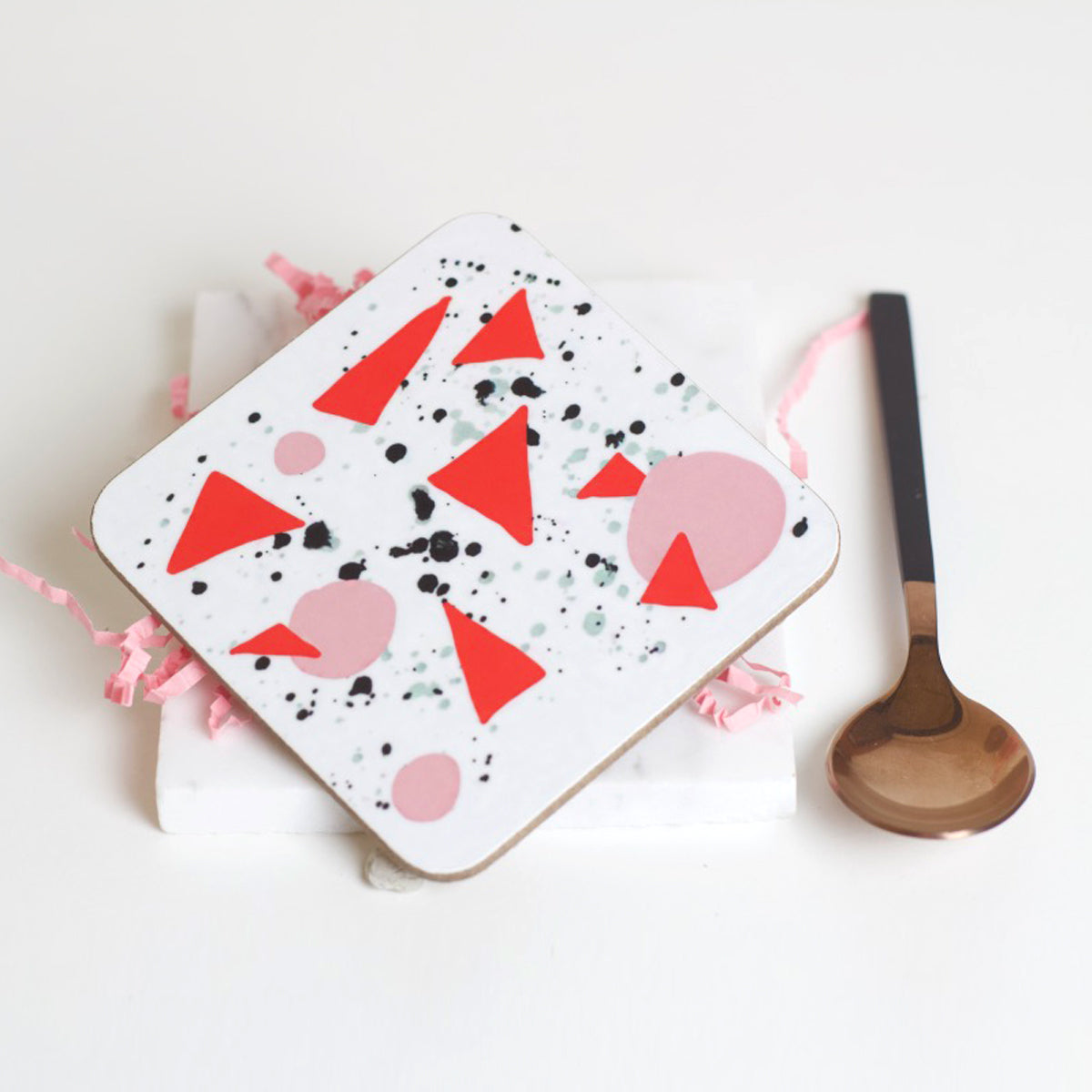 A square coaster with a geometric design consisting of red triangles and pink accents, accompanied by a wooden spoon.
