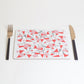 A rectangular placemat with a geometric pattern of red triangles and curvy lines in pink, black, and grey, displayed on a white background, accompanied by a fork and knife.