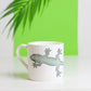 A white bone china mug with a gecko design, placed on a surface with a green background and a palm leaf in the background.