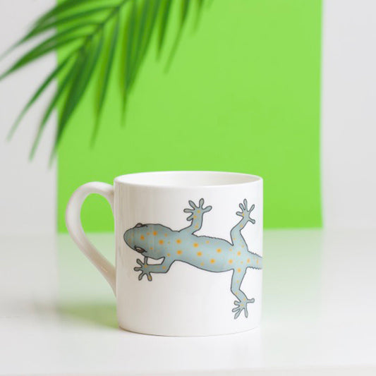 A white bone china mug with a gecko design, placed on a surface with a green background and a palm leaf in the background.