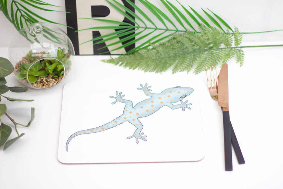 A placemat featuring a drawing of a gecko, with a jungle-themed decoration in the background including a palm leaf and greenery.
