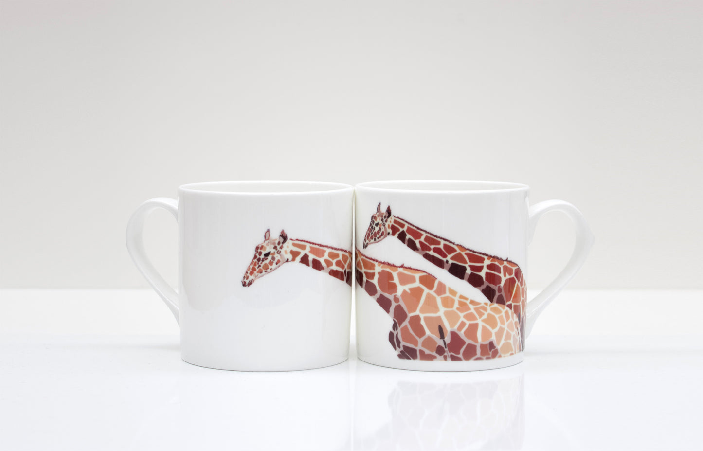 A set of two white bone china mugs with a hand-drawn giraffe design in brown and beige tones, placed on a white surface.