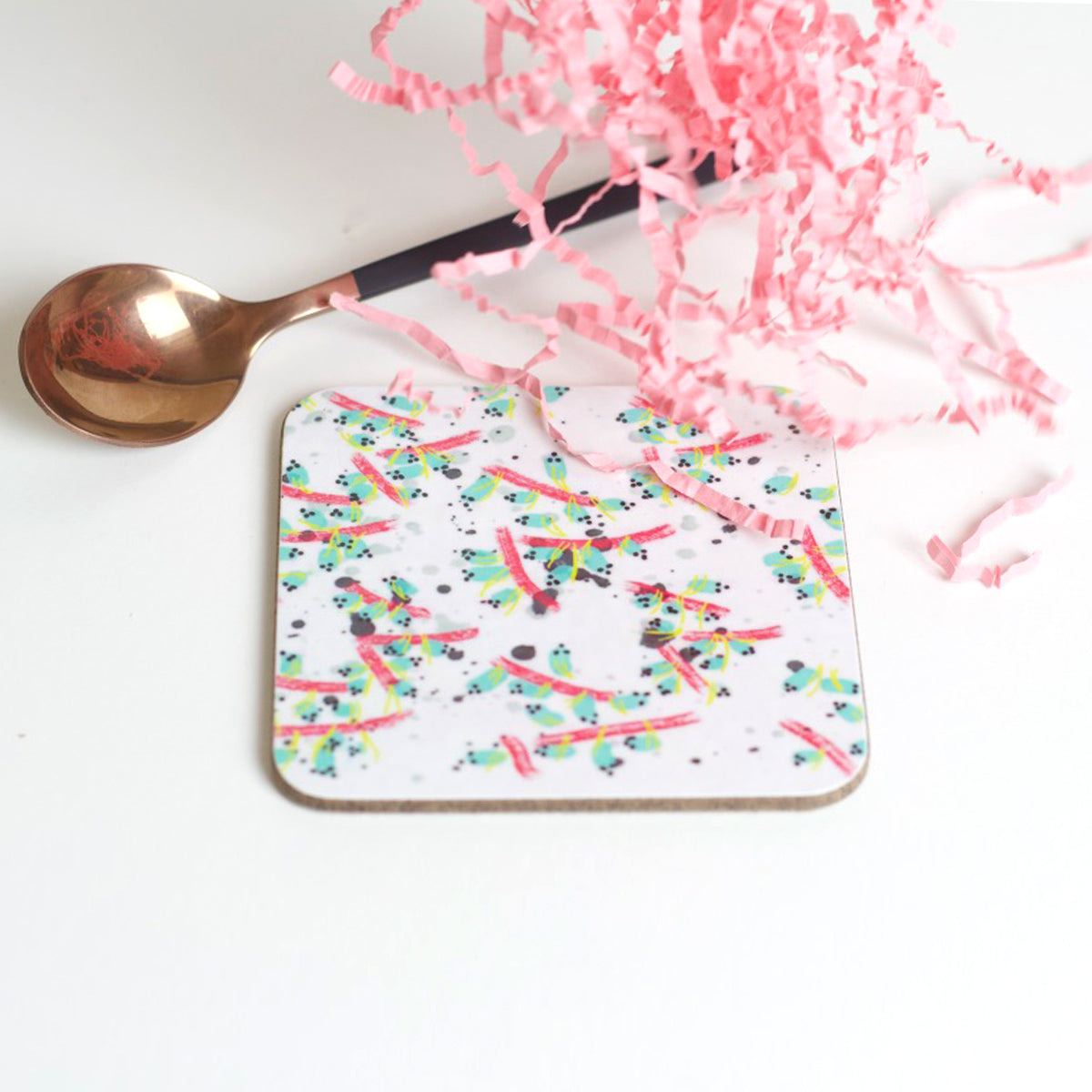 A square coaster with a white background featuring a multicolored, abstract pattern of lines and shapes, placed on a white surface next to a bronze spoon and pink tissue paper.