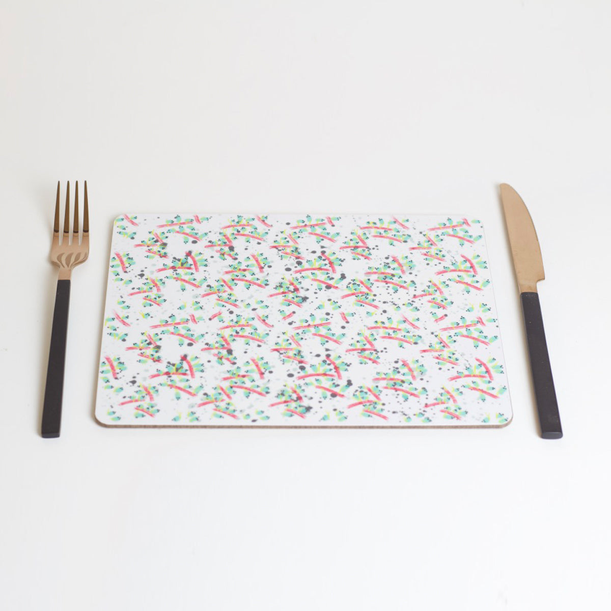 A rectangular placemat with a geometric pattern of overlapping shapes and colors, displayed on a white background. A fork and knife are placed on top of the placemat.