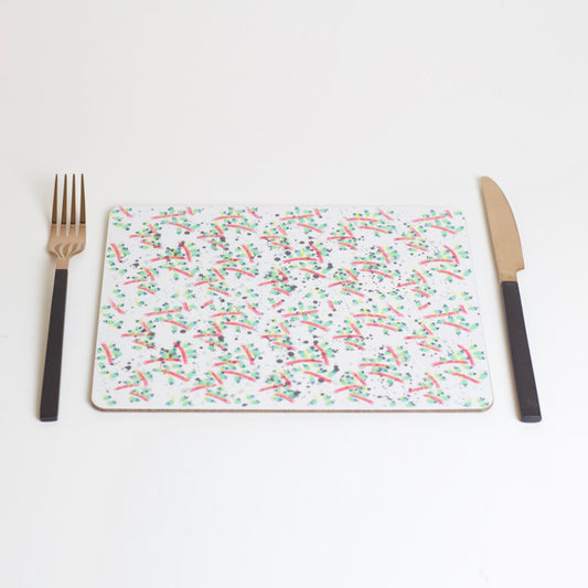 A rectangular placemat with a geometric pattern of overlapping shapes and colors, displayed on a white background. A fork and knife are placed on top of the placemat.