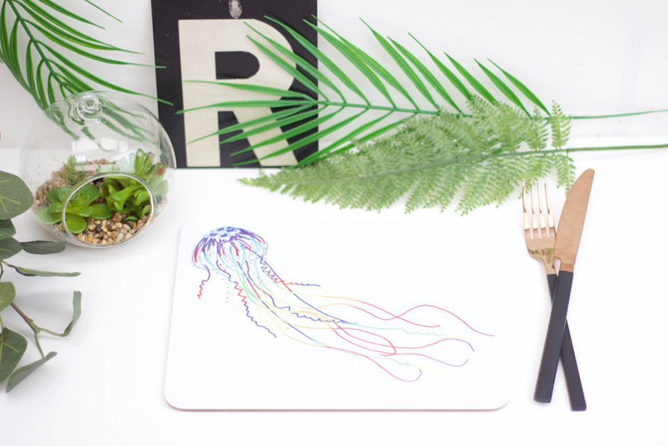 A placemat with a jellyfish design, part of the underwater collection, displayed on a table with a plant, a coaster, and a brush.