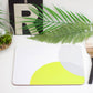 A lime green and grey placemat with a geometric design, overlaid on a white surface, with a wooden cutlery set and a potted plant in the background.
