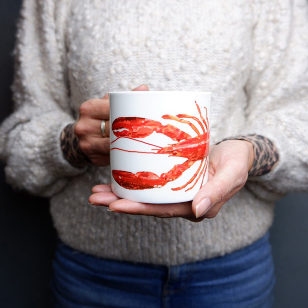 A person holding a white bone china mug with a orange and red lobster design.