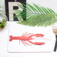 A placemat with a red lobster design, a set of utensils on a white surface with a glass vase with green leaves in the background.