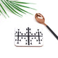 A square hardboard coaster with a black and white geometric symbol design, accompanied by a copper-colored spoon, on a white surface with a green leaf in the background.