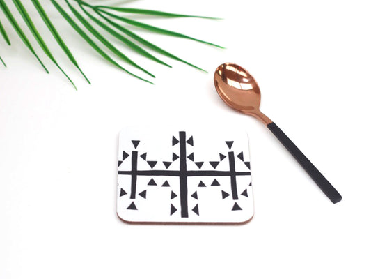 A square hardboard coaster with a black and white geometric symbol design, accompanied by a copper-colored spoon, on a white surface with a green leaf in the background.