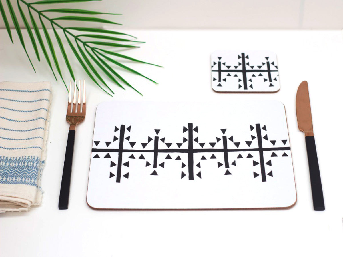 A white and black geometric symbol on a rectangle  placemat which is displayed on a table with a fork, knife, and a napkin. There is a palm leaf in the background.