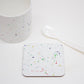 A multicolored splatter coaster with a paint splatter design, placed on a white surface next to a white mug and a white spoon.