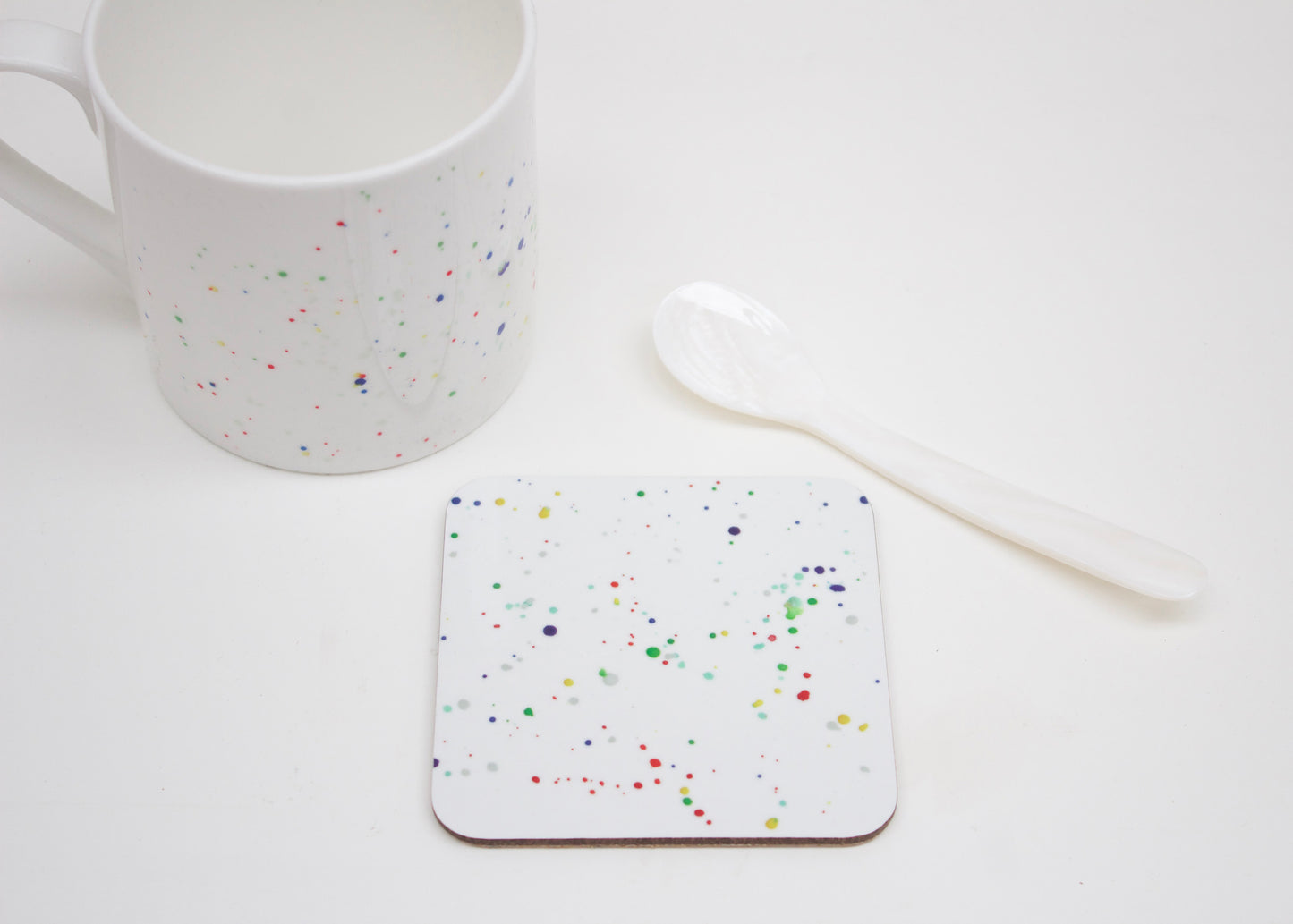 A multicolored splatter coaster with a paint splatter design, placed on a white surface next to a white mug and a white spoon.