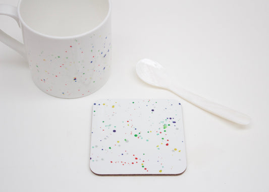 A multicolored splatter coaster with a paint splatter design, placed on a white surface next to a white mug and a white spoon.