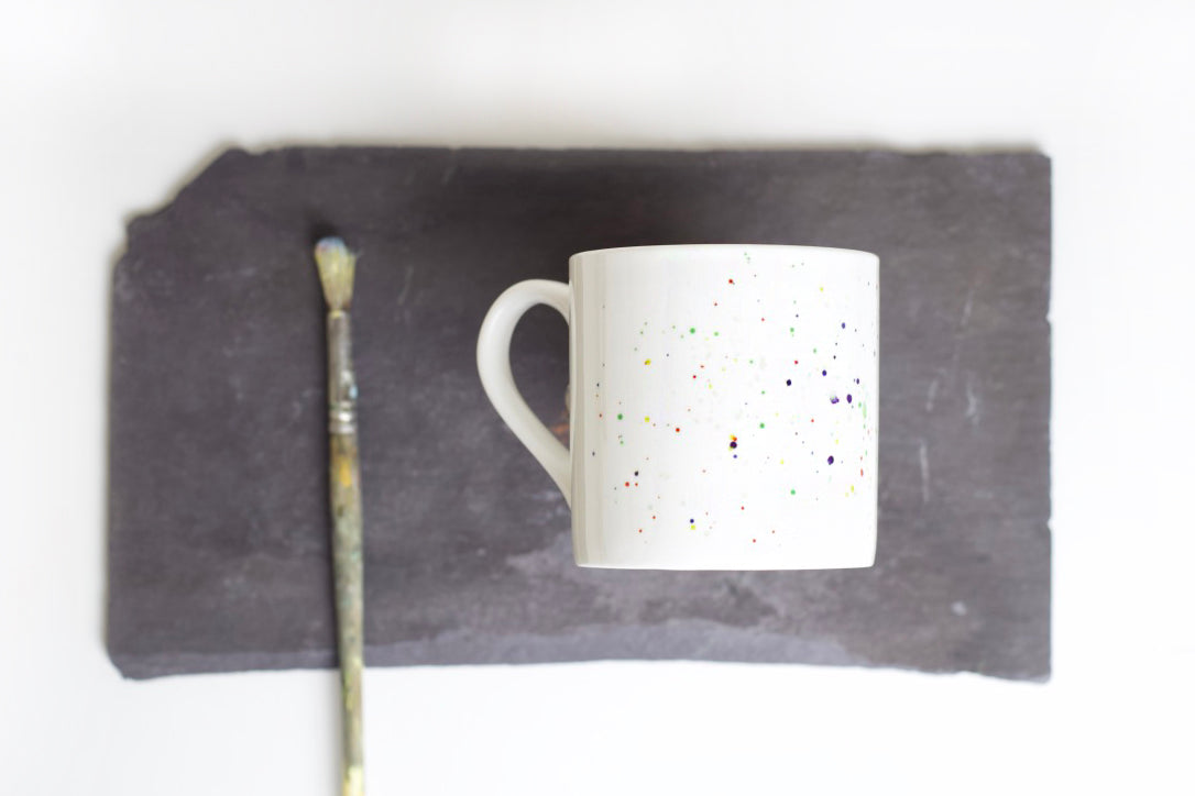 A multicolored splatter pattern bone china mug with a fine rim, displayed on a textured surface, with a brush to the left and a paint mark visible on the mug.