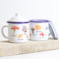 Two enamel mugs with lids, featuring hand-illustrated wild mushroom designs, placed on a wooden board.