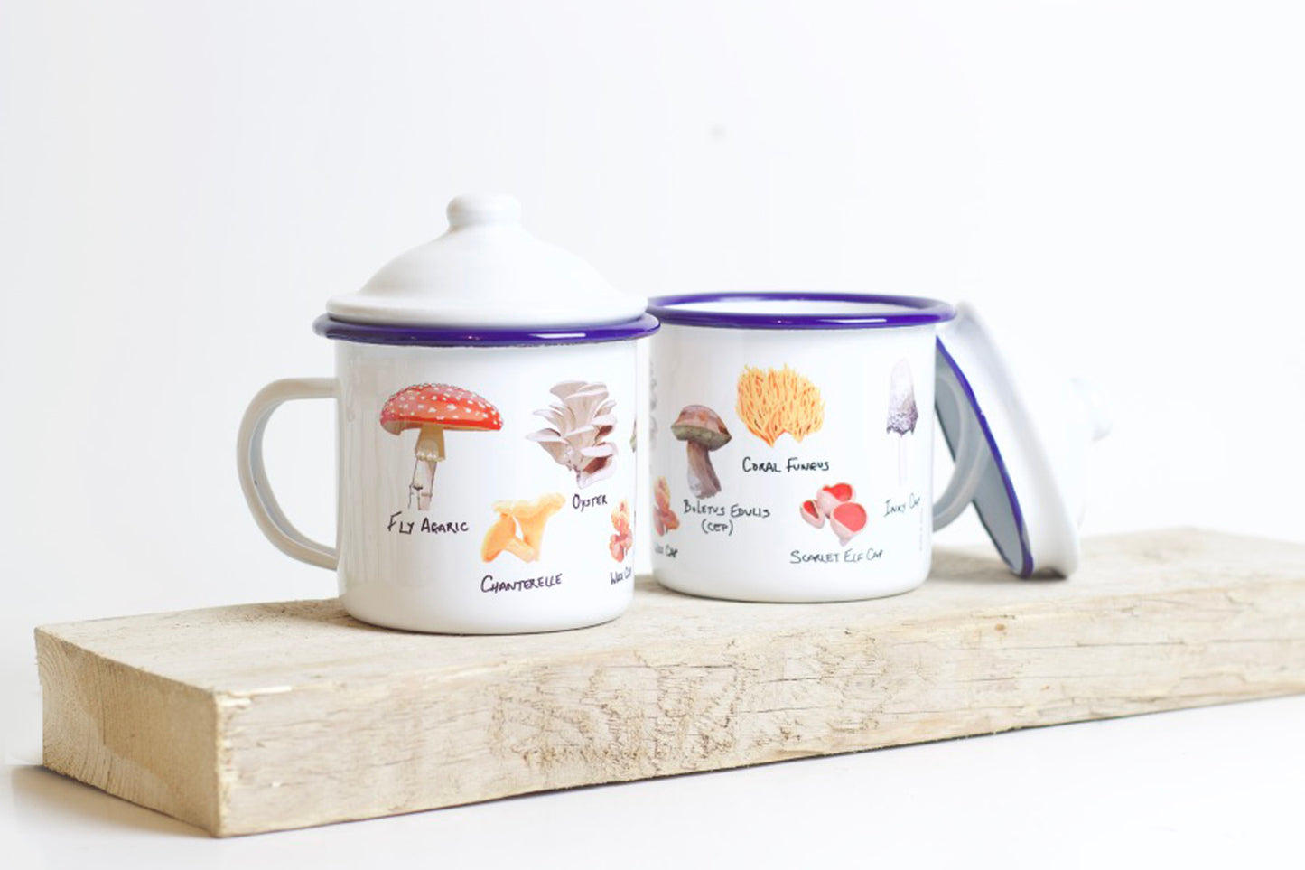 Two enamel mugs with lids, featuring hand-illustrated wild mushroom designs, placed on a wooden board.