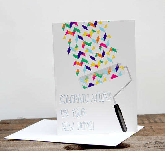 A greeting card with a geometric pattern and the text 'CONGRATULATIONS ON YOUR NEW HOME!' written on it. The card is accompanied by an envelope placed on a wooden surface.