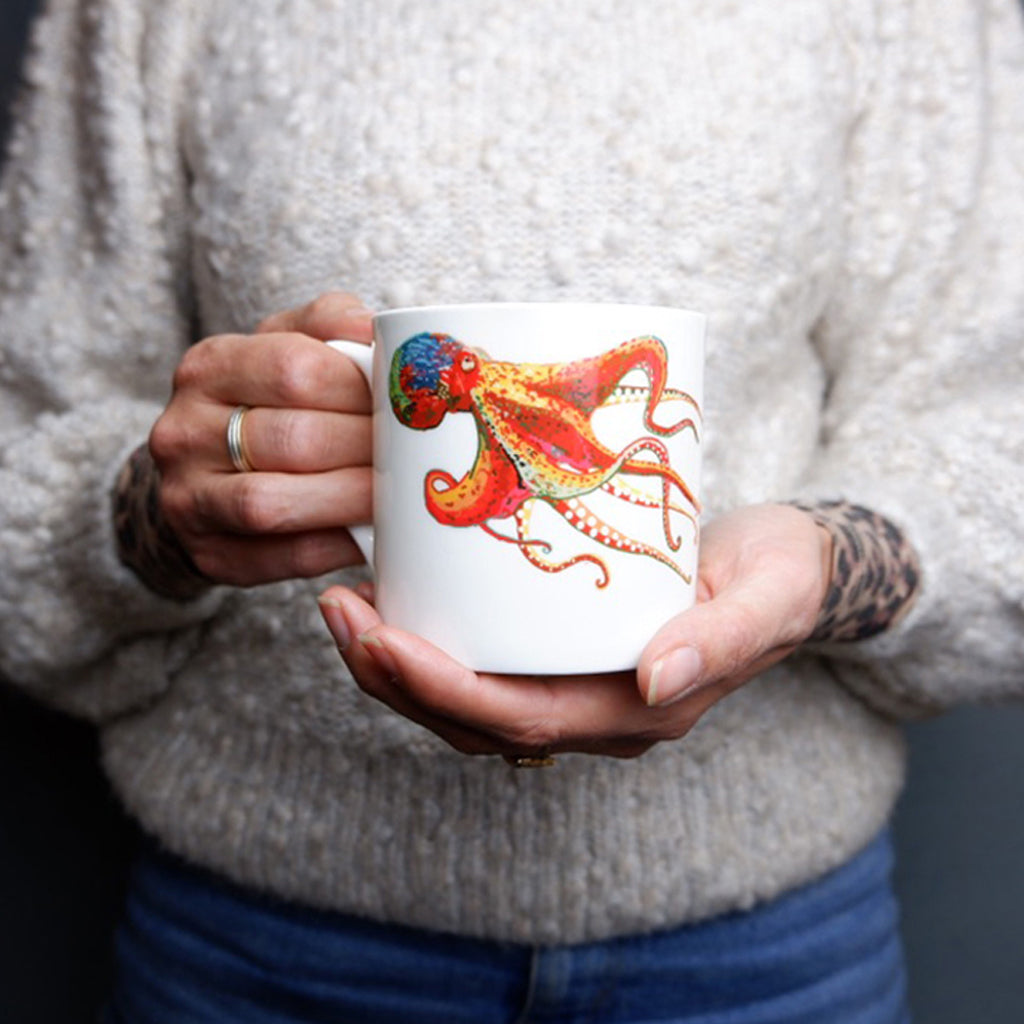 A person holding a white bone china mug with a colorful hand-drawn Octopus design on it.