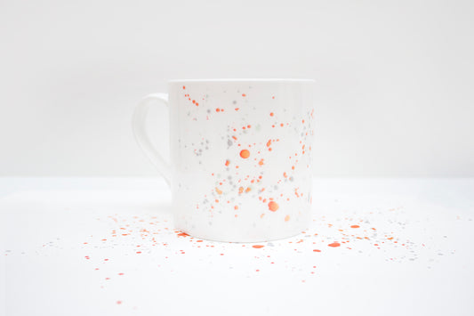 A white bone china mug with orange and grey speckled pattern on a white background, with a fine rim and translucent quality.