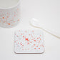 A white coaster with orange splatter design placed on a white surface, with a mug and a white spoon beside it.