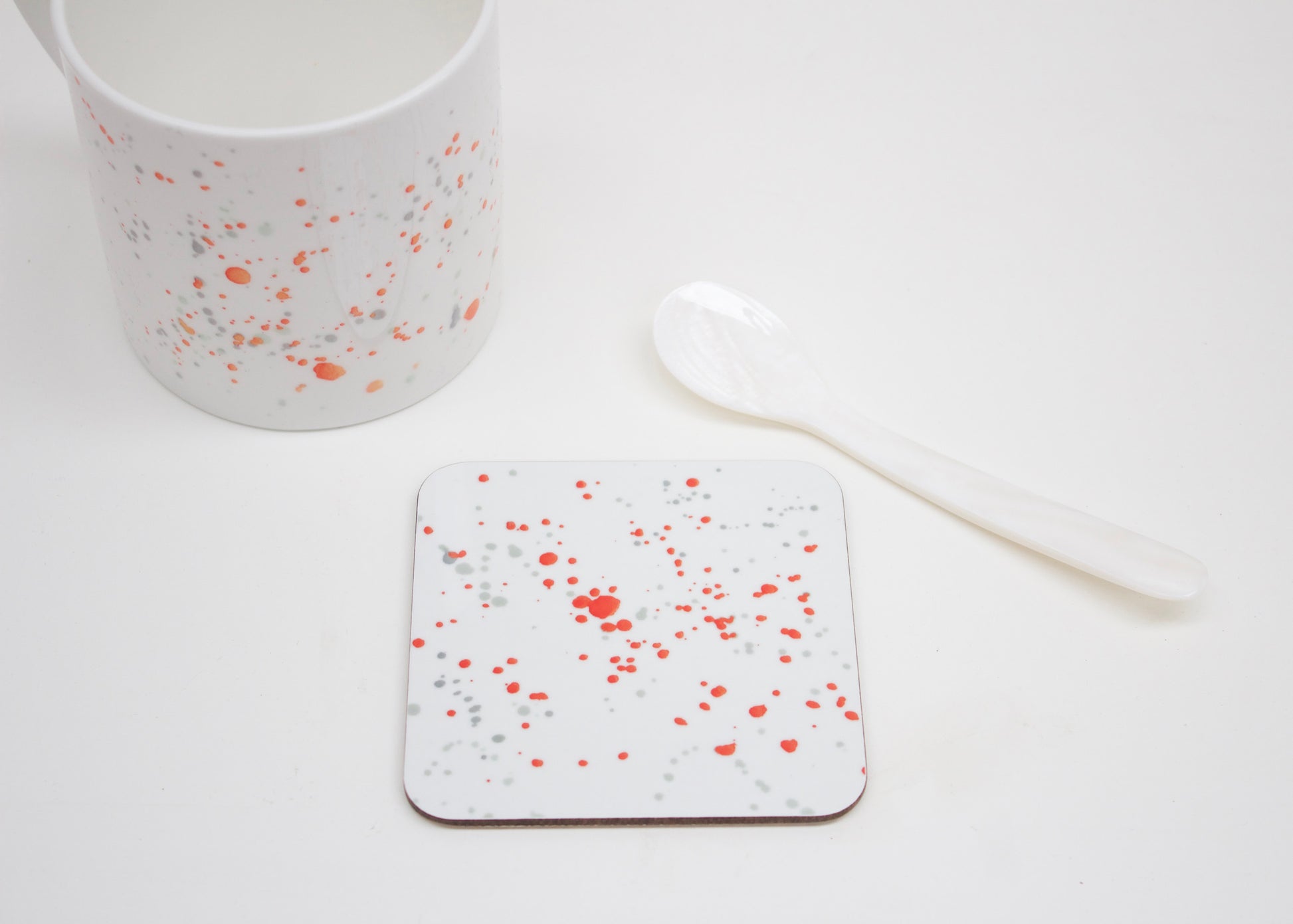 A white coaster with orange splatter design placed on a white surface, with a mug and a white spoon beside it.