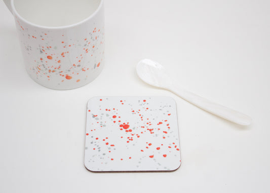 A white coaster with orange splatter design placed on a white surface, with a mug and a white spoon beside it.