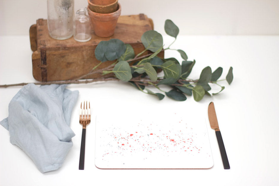 A splatter placemat in orange with multicolored specks, placed on a white surface with a blue napkin, a fork, and a potted plant in the background.