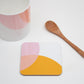 A coaster with a geometric design in orange and pink shades, alongside a bone china mug and a wooden spoon, all placed on a white surface.