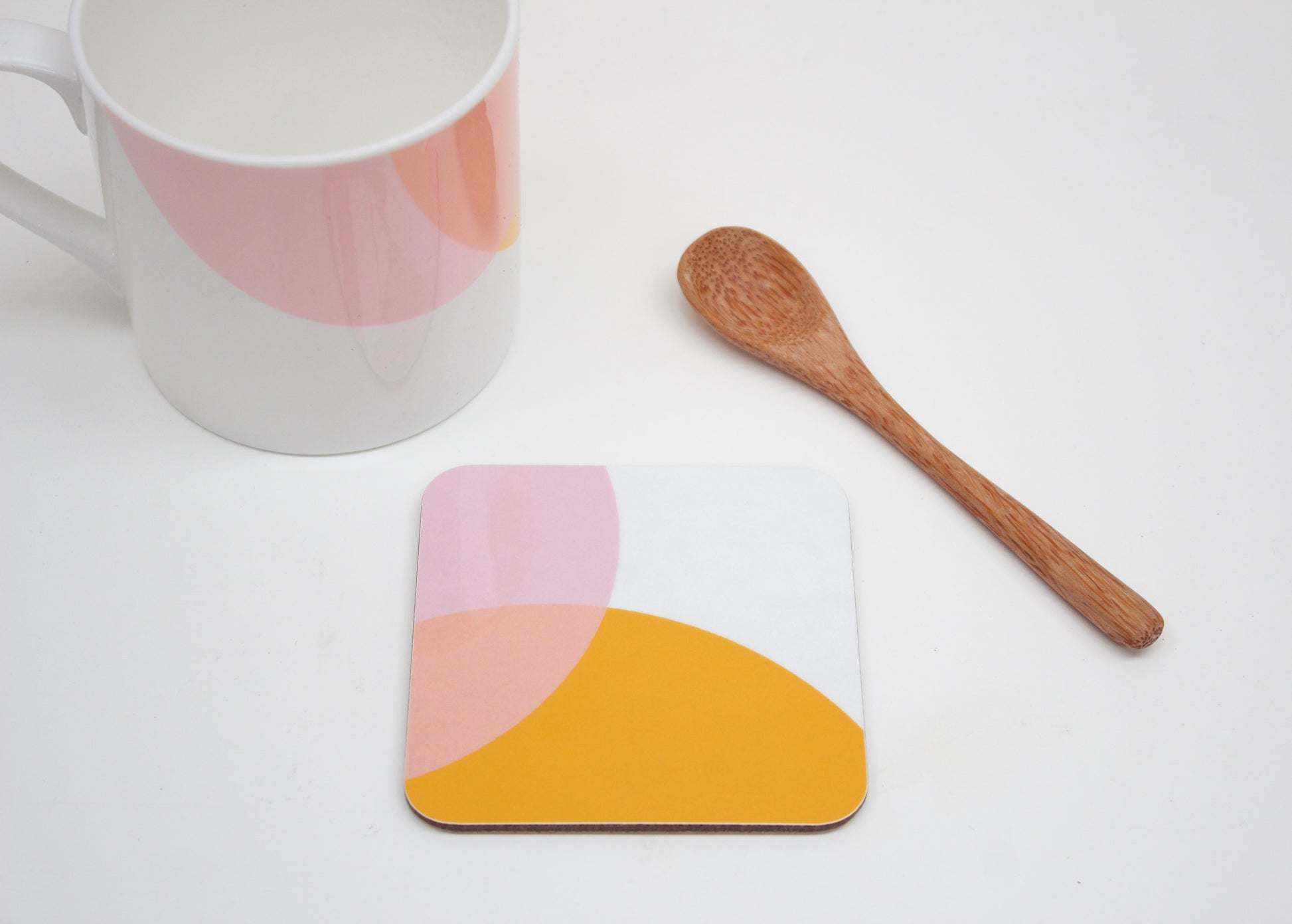 A coaster with a geometric design in orange and pink shades, alongside a bone china mug and a wooden spoon, all placed on a white surface.