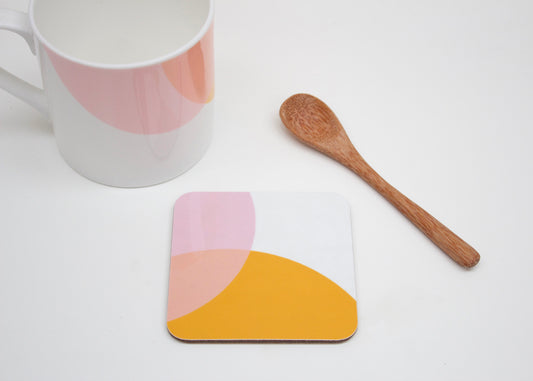 A coaster with a geometric design in orange and pink shades, alongside a bone china mug and a wooden spoon, all placed on a white surface.