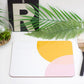 A placemat with a geometric design featuring orange and pink shades, placed on a table with a plant, a glass, and a wooden fork.