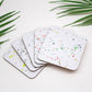A set of four square coasters with a white base and multicolored splatter design, displayed on a white surface.