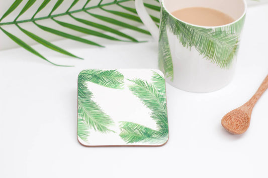 A square coaster with a tropical palm leaf design, placed on a table with a white mug and a wooden spoon, with a real palm leaf in the background.